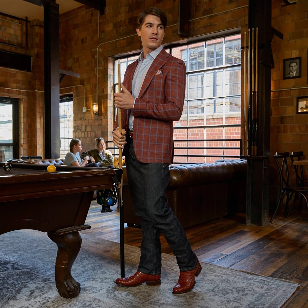 Un hombre que lleva las botas Caiman Tony Lama Tremaine de 12 pulgadas en color marrón, combinadas con jeans oscuros y una chaqueta a cuadros roja, de pie con confianza en un elegante salón con una mesa de billar y paredes de ladrillo rústico al fondo. Compre las botas Tremaine Caiman de 12 pulgadas.